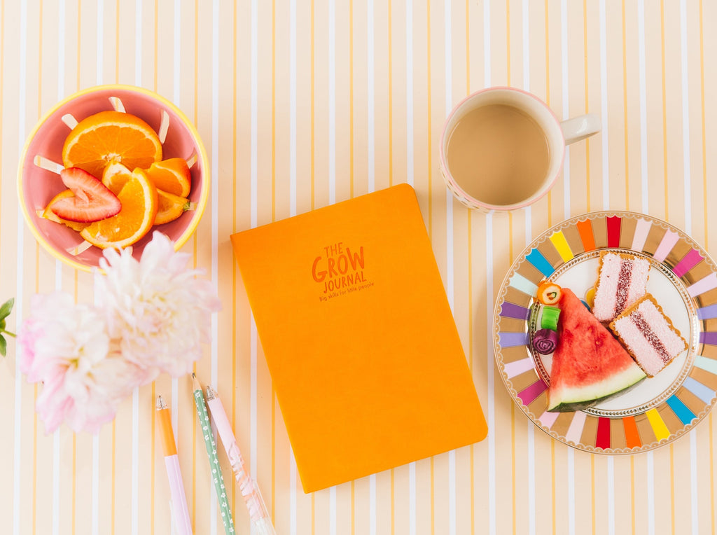 The Grow journal in orange styled with fruit, a cup of tea and stationery on an orange striped background