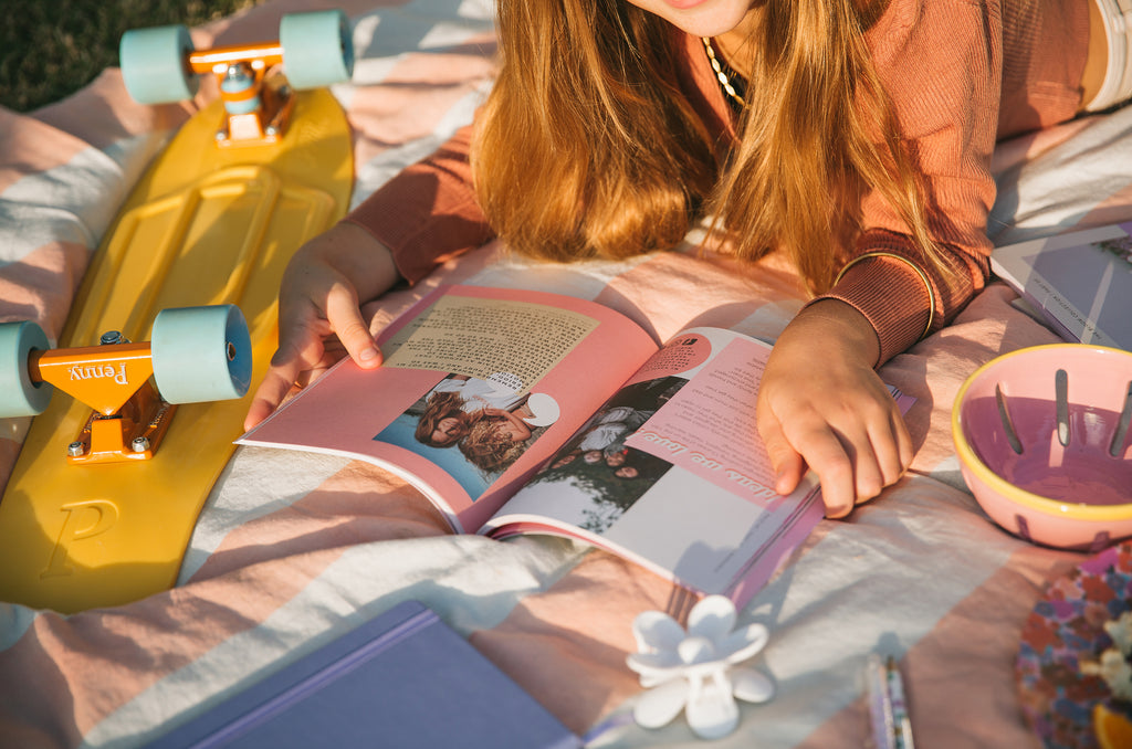 teen girl reading a bloom collection book on a picnic rug. there is a yellow skateboard to her left and stationery in the foreground.